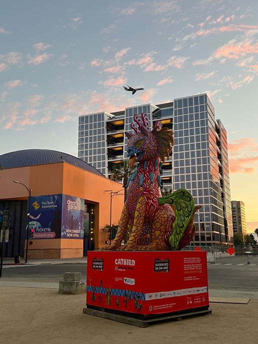 An airplane flies over The Tech Interactive at sunset.  CATBIRD, a beautiful Alebrije statue is in the foreground in La Plaza de Cesar Chavez