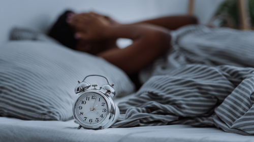 Image of a woman with insomnia lying in bed and covering her face with her hands. An alarm clock shows it is 4am.