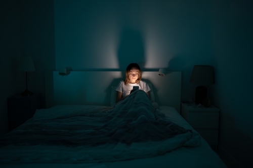 Image in which a woman sits in bed and looks at a smartphone.