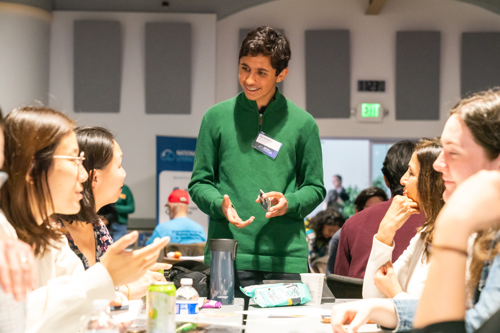 A student in a green sweater with dark hair explains a card game to a group of adults.