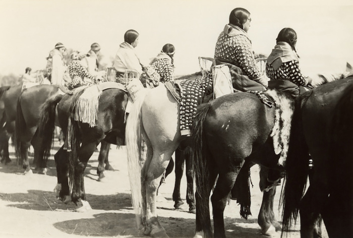 An old black-and-white photo of Native American women and girls in traditional dress riding horses.
