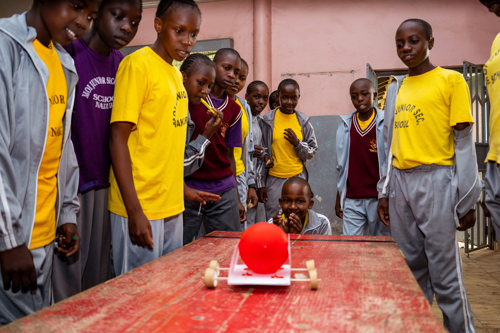 Students watch as they test their device in The Tech Challenge. The device has a platform with four wooden wheels and carries a red ball.