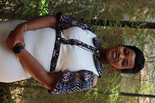 Dr. Shikoh Gitau stands in front of bamboo trees. She is African with short black hair, a big smile, and a white dress.